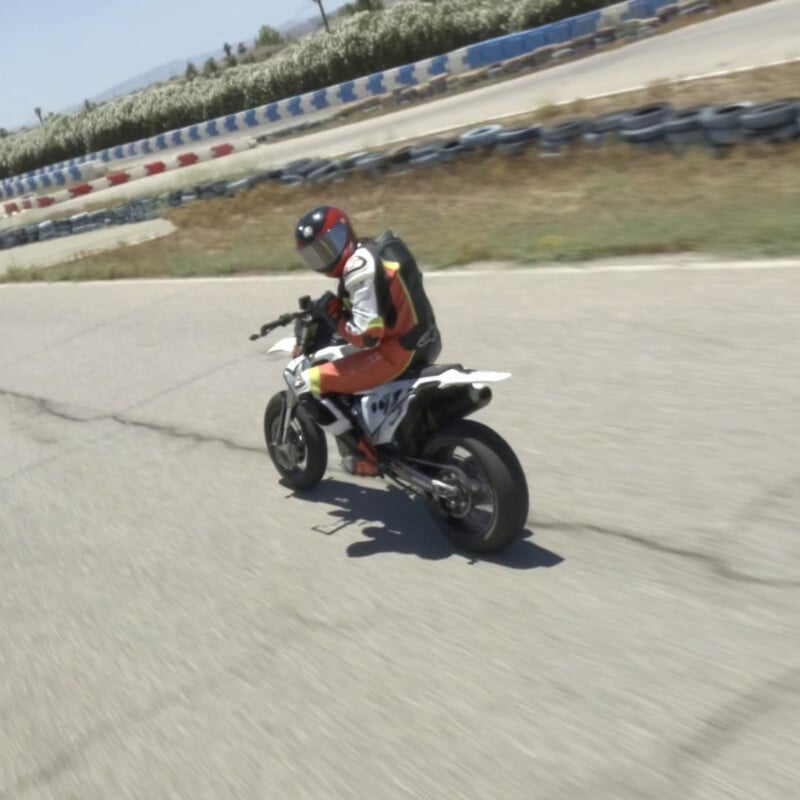 Persoon in rood-wit racepak en helm rijdt op een sportmotor op een circuit, bocht makend. Banden en barrières op de achtergrond.