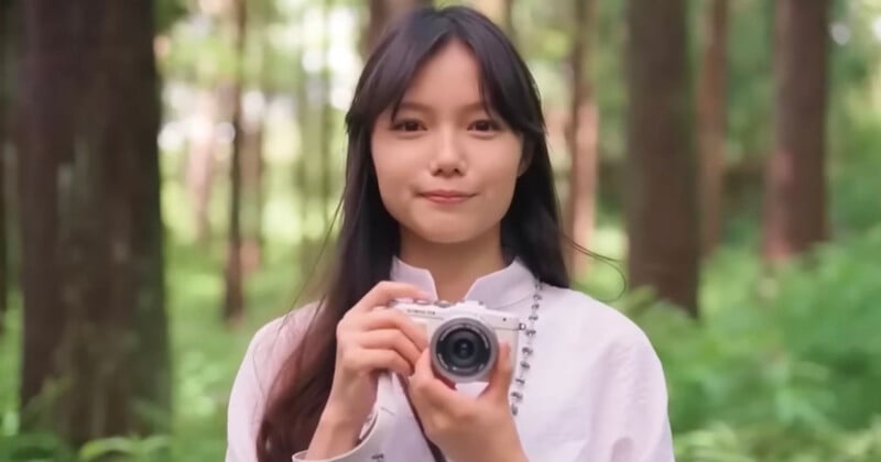 A young woman with long dark hair, wearing a light-colored shirt, stands in a forest holding a camera and smiling softly at the camera. Tall trees and green foliage fill the background.