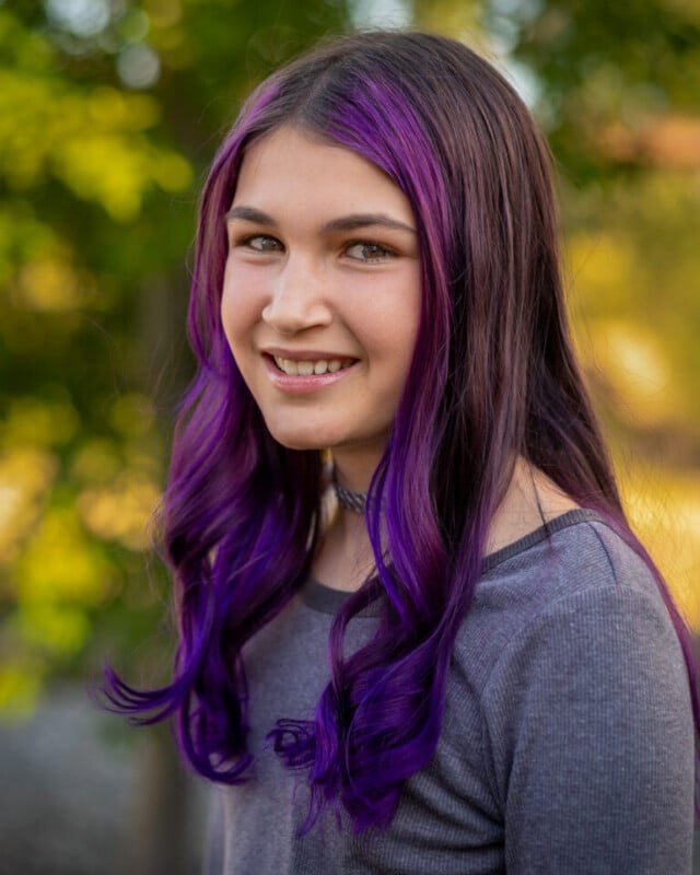 A young person with long, wavy purple hair is smiling and wearing a gray top, standing outdoors with green foliage blurred in the background.