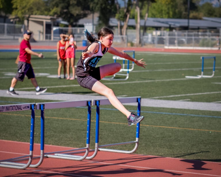 A young girl in athletic gear jumps over a hurdle on a track during a race, with other athletes and people visible in the background on a sunny day.
