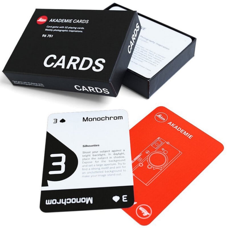 A black box labeled "AKADEMIE CARDS" is open, showing two cards: one black-and-white card titled "Monochrom" with text instructions, and one orange card with a line drawing of a camera.