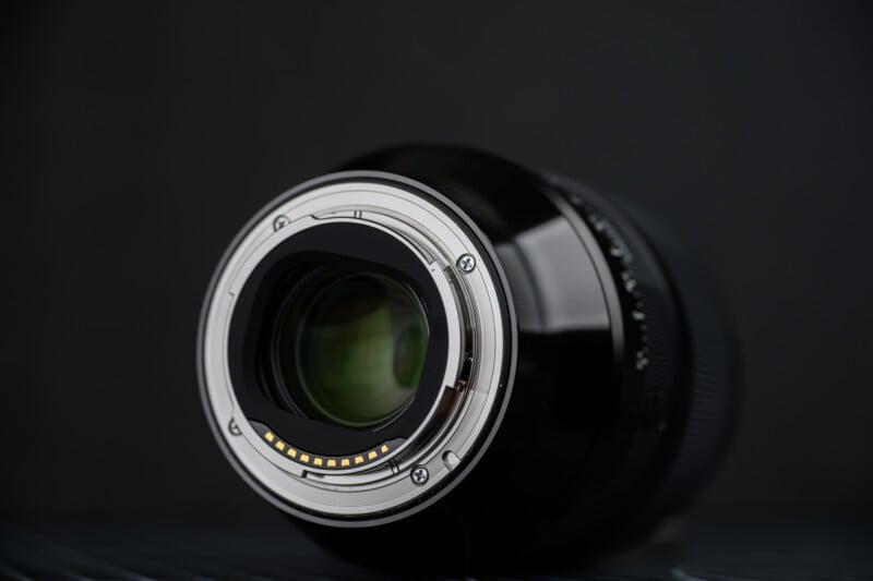 Close-up view of the metal lens mount and glass elements of a camera lens, positioned at an angle against a dark, blurred background.