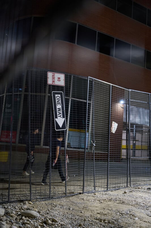 A tilted "One Way" sign and a "No Parking" sign are attached to a tall metal fence at night. Two people walk behind the fence near a building, and the ground is rough with dirt and rocks.