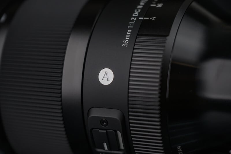 Close-up of a black camera lens showing the focus ring, aperture markings (35mm 1:1.2), and a circular emblem with the letter "A" on it.