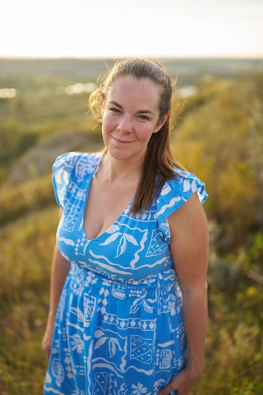 A woman in a blue patterned dress stands outdoors in a grassy field, smiling softly at the camera with sunlight illuminating her and a blurred landscape in the background.