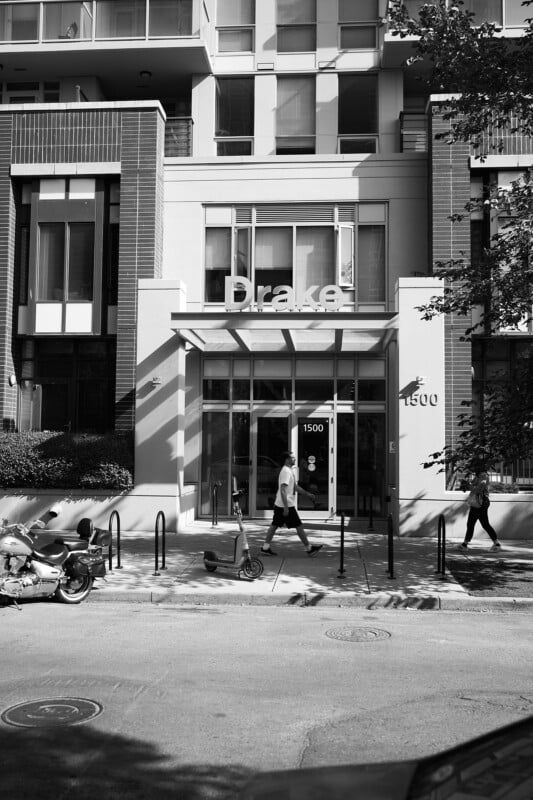 A man walks past the entrance of a modern building labeled "Drake" at 1500, with bike racks, a scooter, and a motorcycle parked outside on a sunny day.