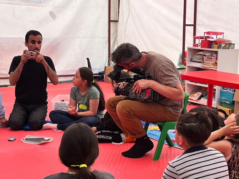 A man films a group of children seated on the floor in a brightly lit room; another man sits facing the group, appearing to show something to the children. Shelves with books and toys are visible in the background.