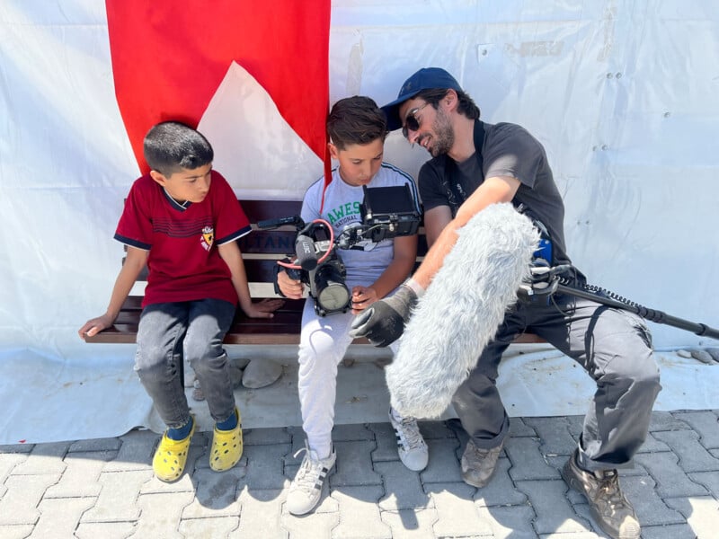 A man with sound equipment helps two boys sitting on a bench, showing them a camera. A large red and white flag hangs behind them. The group appears to be outdoors on a sunny day.