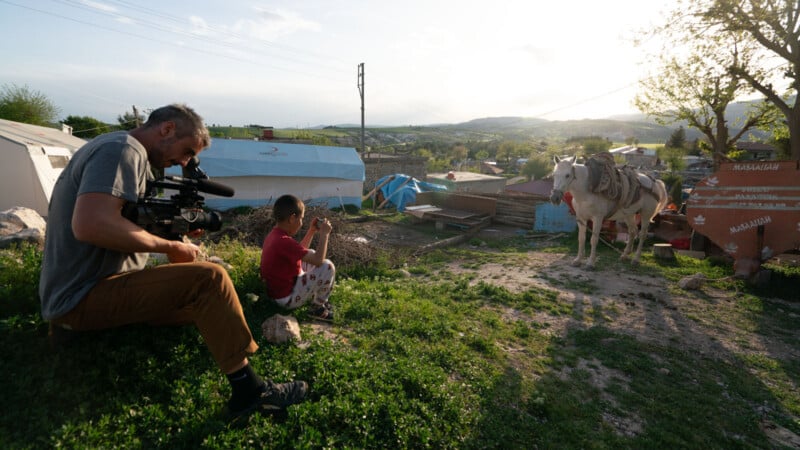 A man with a video camera sits on grass next to a child holding a phone, while a donkey stands nearby, harnessed to a cart, in a rural setting with houses and trees under sunlight.