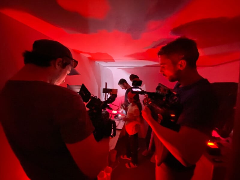 Several people are working in a dimly lit red room, with two individuals in the foreground holding video cameras, filming or preparing to film others further inside the room.