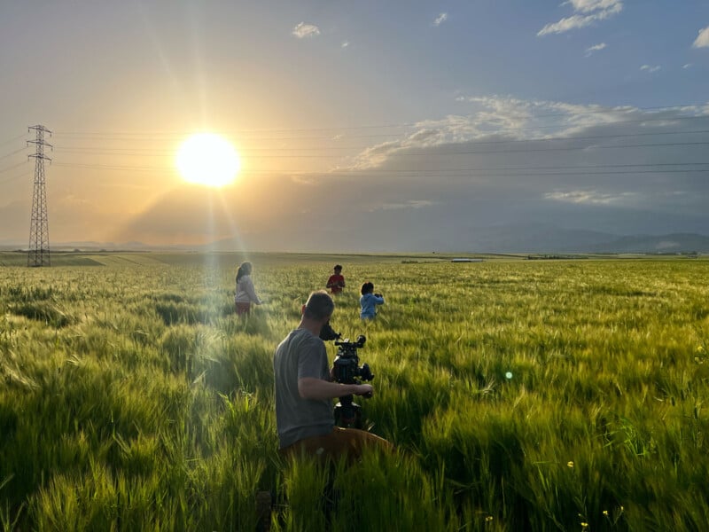 A person with a camera films three children running through a lush green field at sunset, with mountains and power lines visible in the background under a partly cloudy sky.