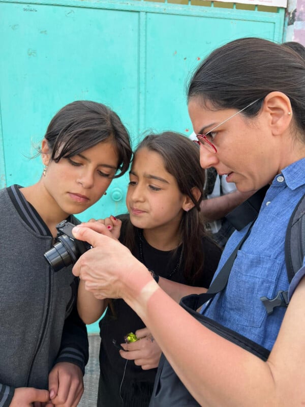 Three people, including two girls and an adult woman, closely look at the screen of a camera the woman is holding. They stand in front of a teal gate, appearing focused and curious.