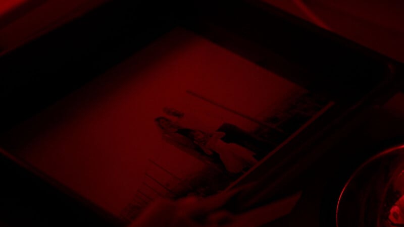 A black-and-white photograph develops in a tray under red darkroom lighting, showing three people posing outdoors.