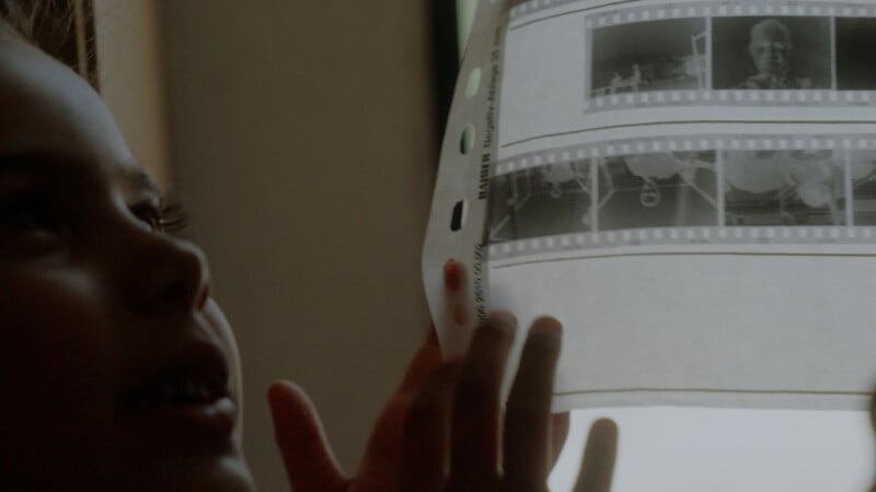 A child holds up a strip of photographic negatives to the light, examining the images closely. The child's face is partially visible in profile, with soft lighting highlighting their features.