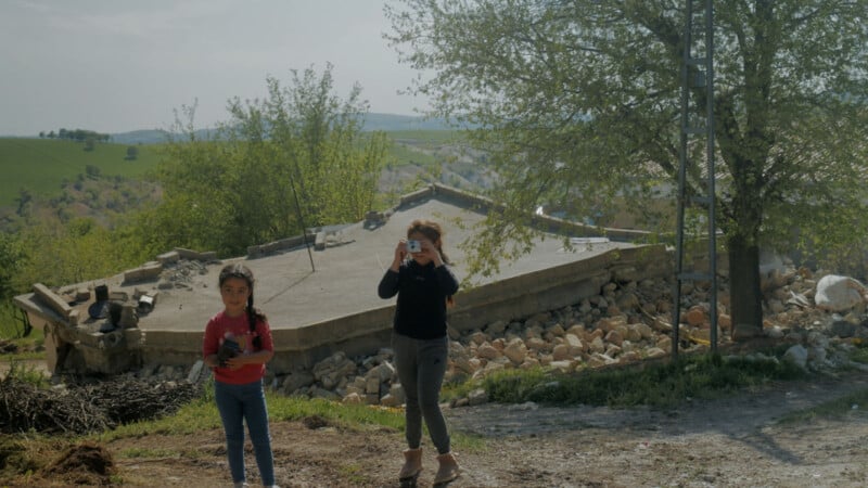 Two young girls stand outside on a dirt path, one holding a camera and taking a photo, the other holding a small object. Behind them is a collapsed house surrounded by rubble and trees, with green hills in the background.