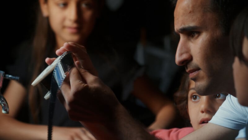 A man closely examines a strip of film while two children watch attentively, with another child and a girl blurred in the background. The scene is warmly lit and focused on the man's careful inspection.