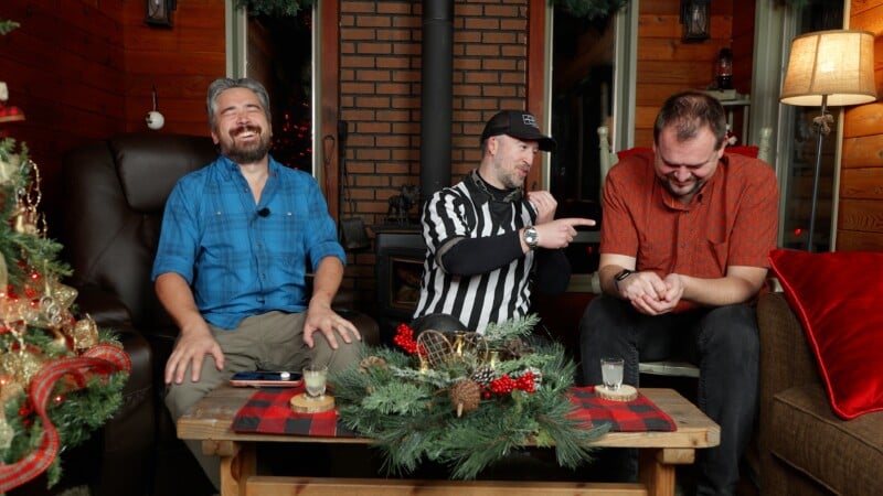 Three men sit on a cozy living room couch decorated for Christmas. The man in the middle wears a black-and-white referee shirt and points while the two others laugh. A festive centerpiece is on the wooden table in front of them.