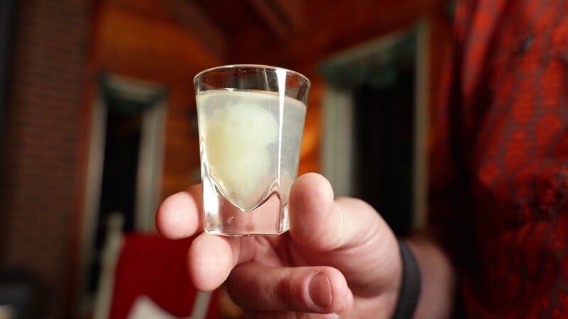 A close-up of a hand holding a shot glass filled with a clear liquid and a floating, cloudy white object, possibly an egg or similar item, with a blurred indoor background.