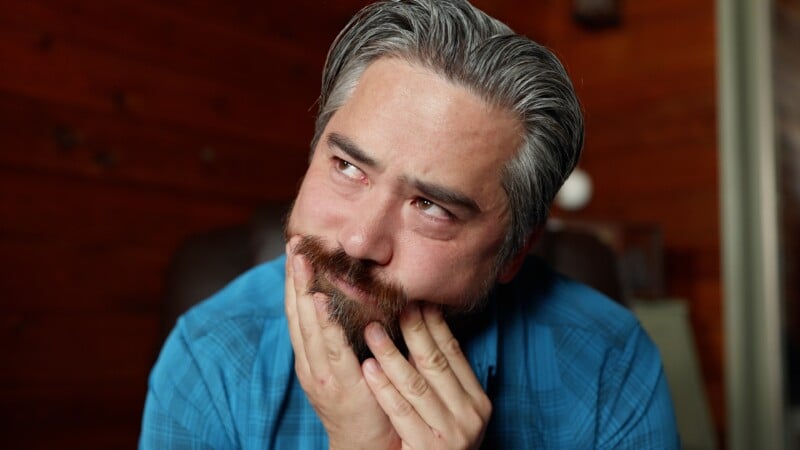 A man with gray hair and a beard, wearing a blue shirt, rests his face in his hands and looks upward with teary eyes, appearing thoughtful or emotional. The background is warmly lit with wooden walls.