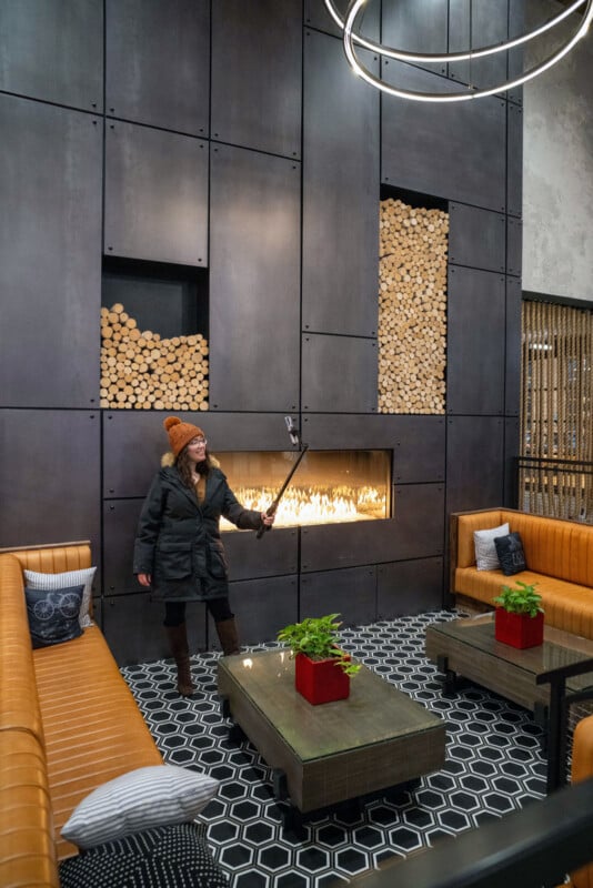 A person in a winter coat and hat takes a selfie in a modern lounge with tan sofas, a glass fireplace, stacked firewood, and geometric black and white flooring. Green plants in red pots sit on coffee tables.