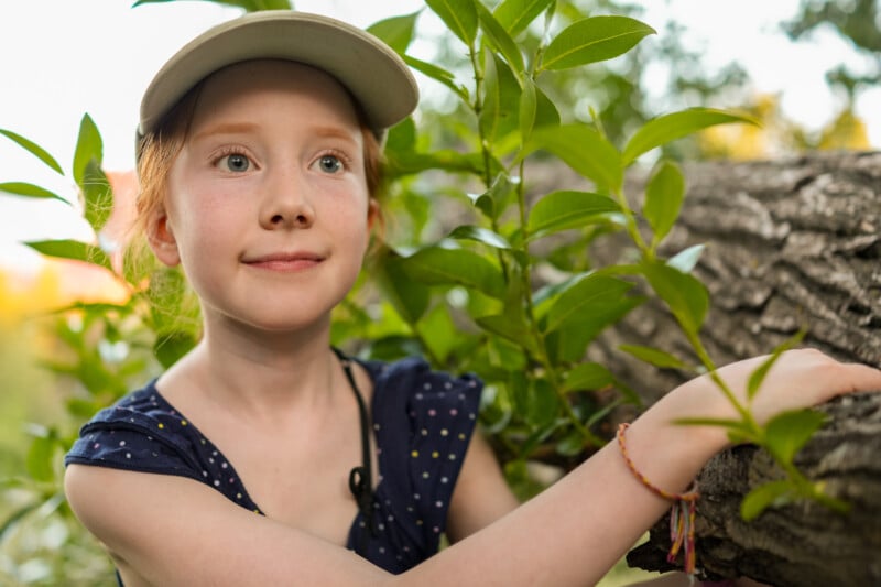 A young girl wearing a light cap and a navy blue polka-dot dress smiles while climbing a tree, surrounded by green leaves and sunlight.