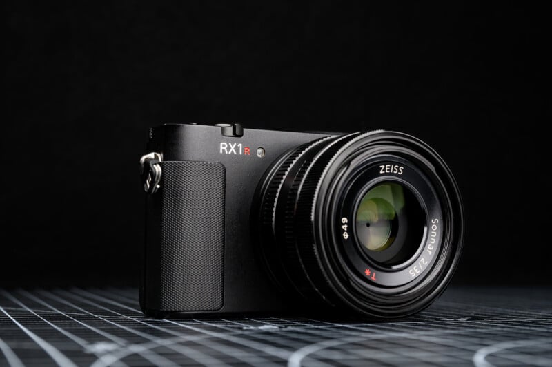 A black Sony RX1R II digital camera with a Zeiss lens is displayed on a grid-patterned surface against a dark background.