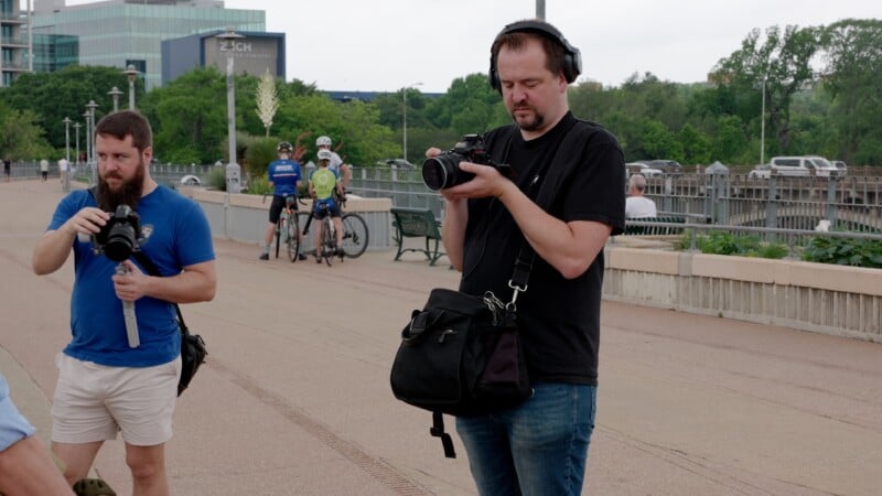 Two men stand on a wide pedestrian path; one wears headphones and holds a camera, the other holds video equipment. Bicyclists and greenery are visible in the background.
