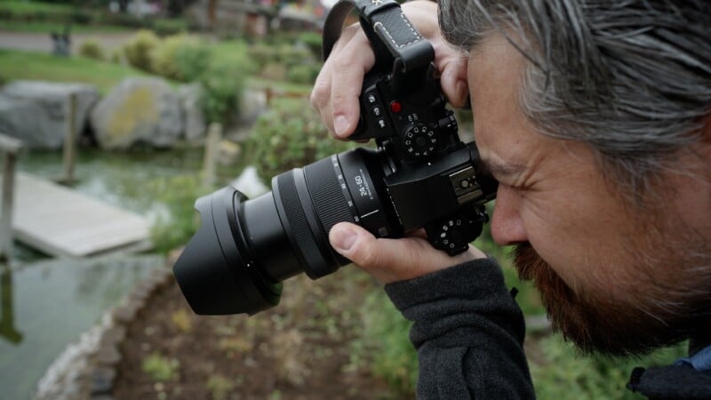 A person with gray hair and a beard is holding a camera up to their eye, focusing intently on taking a photograph outdoors near a pond and a wooden dock.