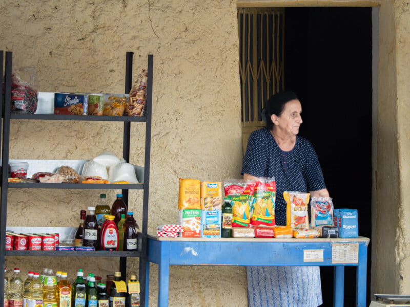 A woman stands behind a small blue table displaying packaged food and goods outside a building with a rough, beige wall. Shelves with bottles and snacks are next to her. She looks to her left, appearing thoughtful.