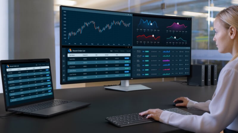 A woman sits at a desk working on a computer with multiple large monitors displaying financial charts, data tables, and graphs in a modern office setting.
