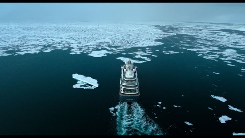 A large ship cruises through dark, icy waters, leaving a wake behind as it navigates through floating ice sheets and scattered icebergs under a gray sky.