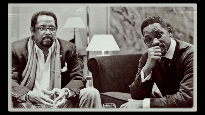 Two men sit indoors on a sofa and armchair, one with glasses and a scarf, the other with his hand on his chin. Both look toward the camera. The setting appears formal, with lamps and a marble wall in the background.