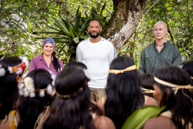 Three people stand facing a group of Indigenous people outdoors. The group members have traditional attire and headbands. Trees and greenery fill the background. The three people in front are smiling or neutral.