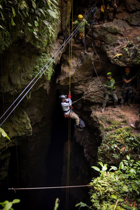 A person wearing a helmet and harness descends on a rope into a large cave, while several others in safety gear stand on the rocky edge above, surrounded by lush green vegetation.