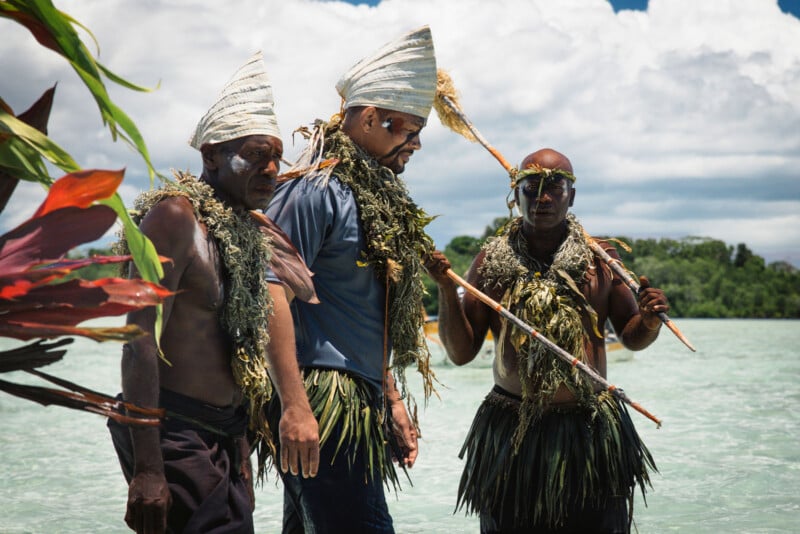 Three men in traditional attire made of leaves and grass, with face paint and headdresses, stand in shallow water under a partly cloudy sky, surrounded by lush greenery and tropical plants.