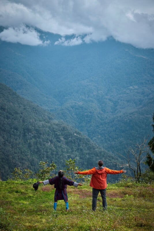 Two people stand on a grassy hill with arms outstretched, facing a scenic view of lush green mountains and cloudy skies in the distance.