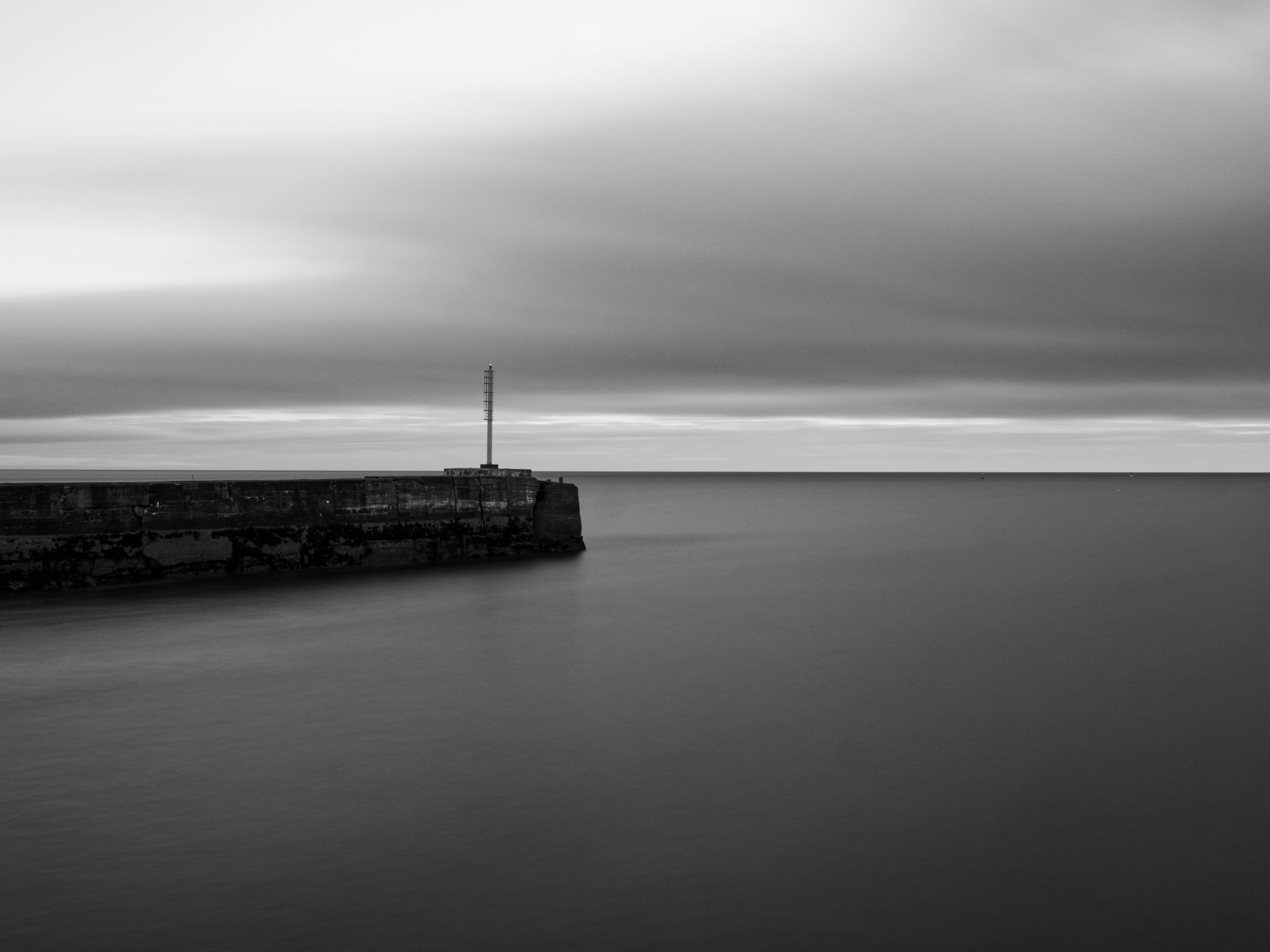 Black and white photo of a calm sea with a concrete pier extending from the left, topped with a tall, narrow pole or mast. The sky is cloudy, and the horizon is visible in the distance.