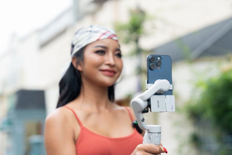 A woman wearing a headscarf and orange tank top smiles while holding a smartphone on a handheld stabilizer, filming outdoors with blurred buildings and greenery in the background.