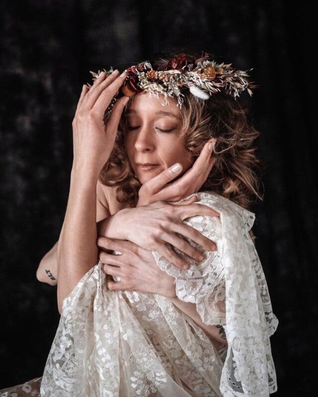 A woman with curly hair and a floral crown closes her eyes, gently touching her face and chest. She is draped in sheer, white lace fabric against a dark background, creating an intimate, serene mood.