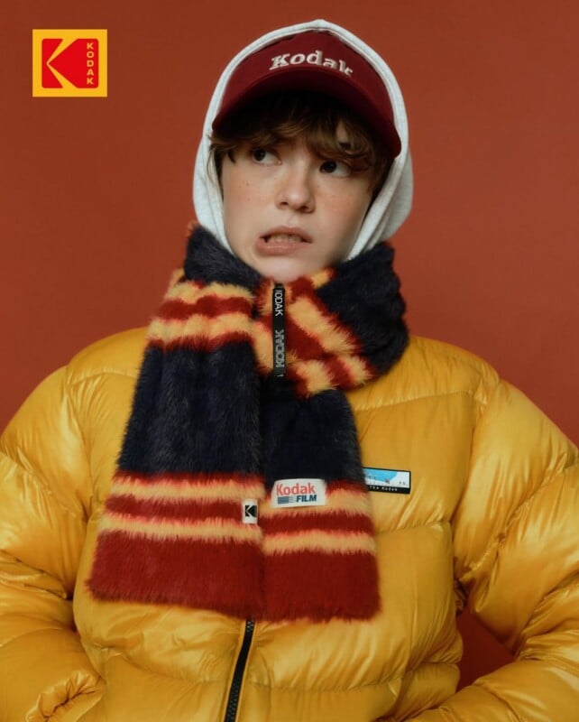 A person wearing a yellow puffy jacket, a maroon and yellow striped scarf, and a maroon Kodak cap stands against a red background with a Kodak logo in the top left corner.