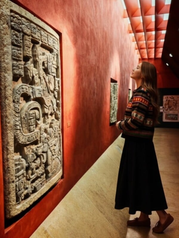 A woman in a colorful sweater and black skirt stands in a museum, observing an ancient stone relief with intricate carvings on a red wall under geometric ceiling lights.