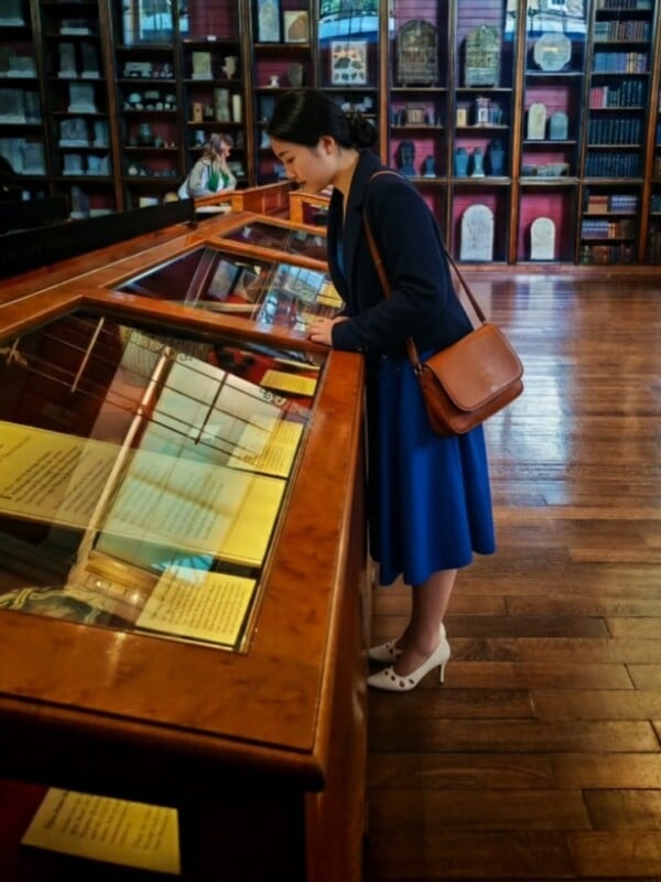 A woman in a blue dress and white heels reads documents in a glass display case at a museum or library, with shelves of books and artifacts in the background.