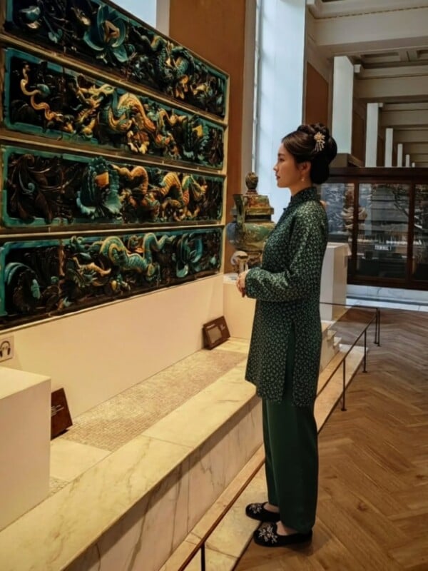 A woman in a green traditional outfit stands in a museum, observing ornate, colorful ceramic reliefs mounted on the wall. The gallery features wooden floors, glass displays, and sculptures in the background.