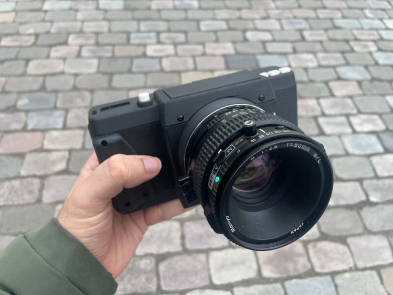 A person holding a black digital camera with a large lens, standing on a cobblestone surface with rectangular gray and beige stones.
