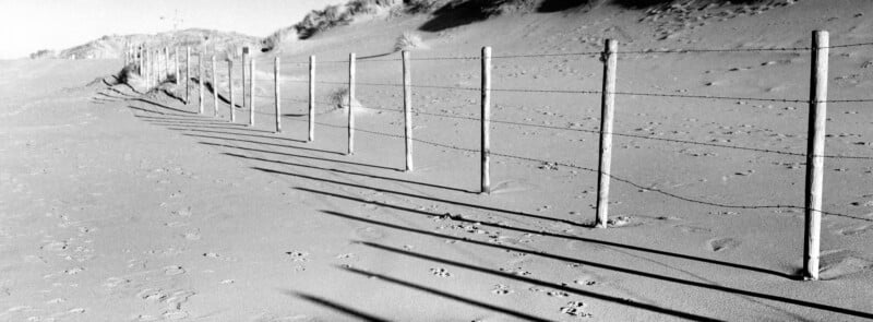 A barbed wire fence casts long, sharp shadows on sandy dunes with footprints and grass-covered hill in the background, in a black-and-white landscape scene.