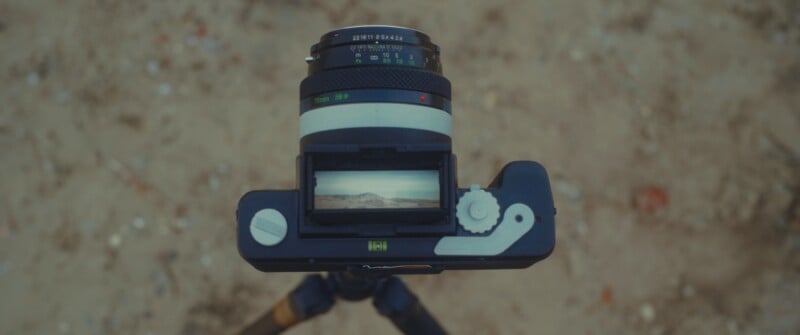 A close-up, top-down view of a vintage camera on a tripod, with its viewfinder displaying a landscape scene featuring distant mountains and a cloudy sky. The background is blurred, showing sandy ground.