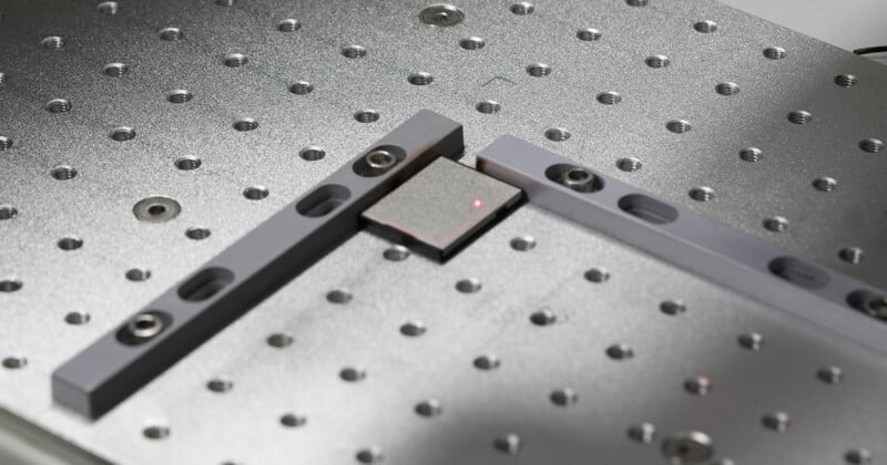 A close-up of a metallic surface with a grid of holes, featuring two gray metal bars clamping a small rectangular object with a red laser dot on it.