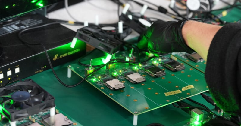 A gloved hand adjusts cables on a green circuit board with glowing green lights and multiple components, surrounded by fans and a KVM switch on a workbench.