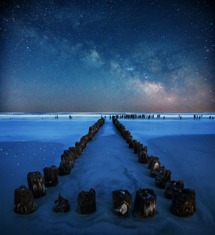 Old wooden posts form two parallel lines on a sandy beach at night, leading toward the ocean. The sky is filled with stars and the Milky Way, creating a serene and mystical atmosphere.