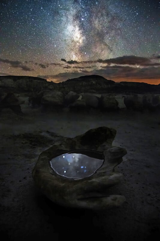 A night scene shows the Milky Way in a star-filled sky above rocky hills. In the foreground, a large rock shaped like a cupped hand holds water, reflecting stars and part of the sky above.